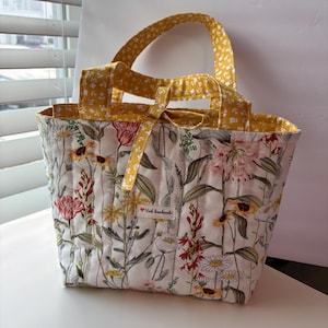 May include: A quilted tote bag featuring a white fabric base with a colorful botanical print of wildflowers in shades of red, yellow, and green. The bag has yellow handles with a small white floral pattern and a small label reading Vied handmade.