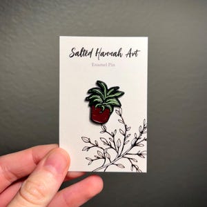 May include: An enamel pin featuring a potted plant with green leaves and a red pot. The pin is on a white card with the text "Salted Hannah Art" and "Enamel Pin". A black and white illustration of a plant branch is also on the card.