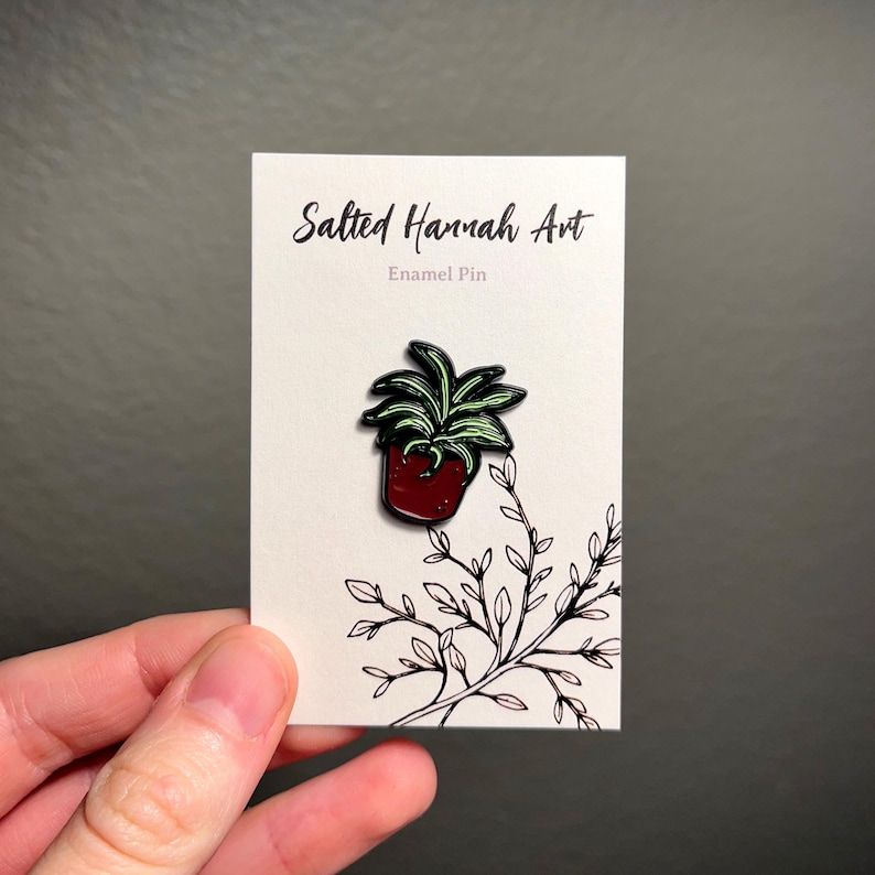 May include: An enamel pin featuring a potted plant with green leaves and a red pot. The pin is on a white card with the text "Salted Hannah Art" and "Enamel Pin". A black and white illustration of a plant branch is also on the card.