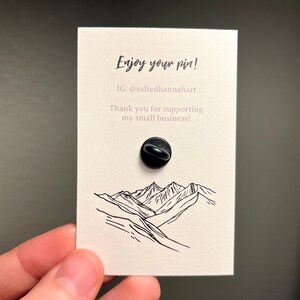 May include: A black enamel pin on a cream-colored card with the text "Enjoy your pin!" and "Thank you for supporting my small business!" The card features a mountain range illustration.