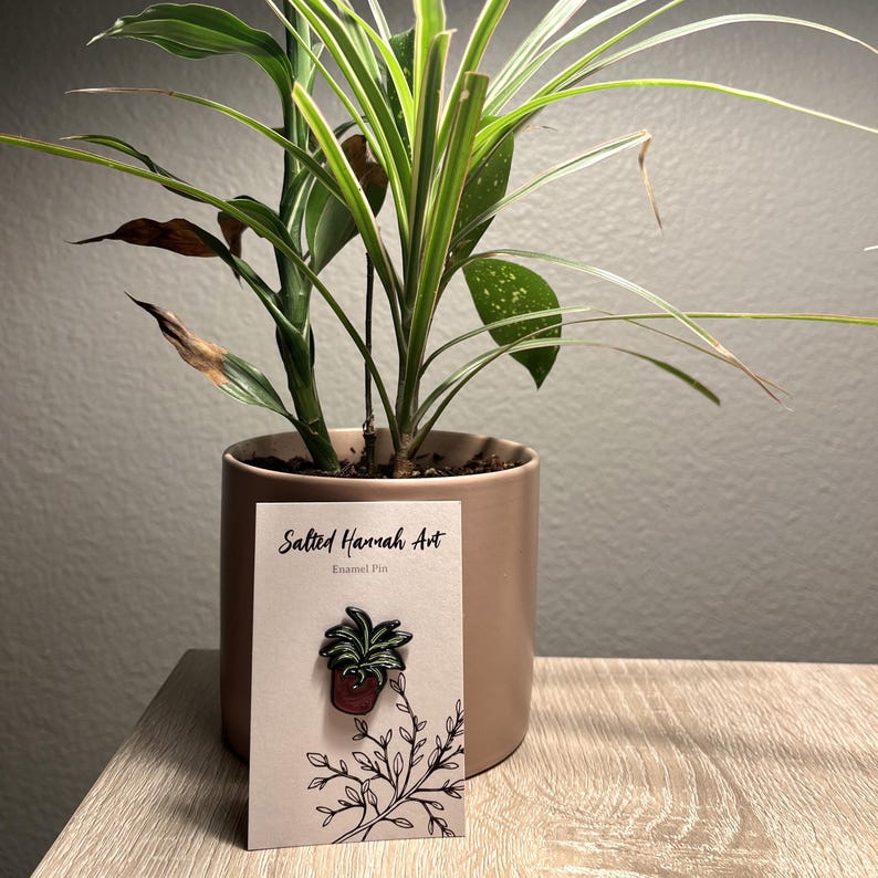 May include: An enamel pin featuring a potted plant design, displayed on a card with the text "Salted Hannah Art." The pin is set against a backdrop of a live plant in a terracotta pot, placed on a wooden surface.