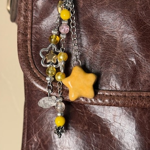 Puede incluir: Un llavero decorativo con temática de estrellas. El llavero presenta un gran dije de estrella amarilla, un dije plateado en forma de estrella y varias cuentas en colores amarillo, rosa y transparente. El llavero está sujeto a una cadena y un anillo plateados.