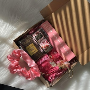 Peut inclure: Une boîte cadeau marron remplie de produits de beauté, dont du parfum, un chouchou et un produit pour les lèvres. La boîte contient un flacon de parfum Tom Ford, un flacon de parfum Gucci Flora et un produit pour les lèvres rose avec le texte "SUMMER SUNDAY".