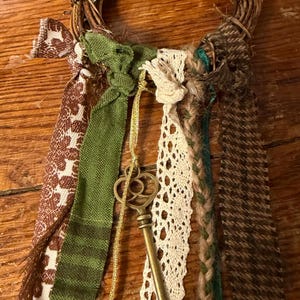 May include: A rustic wreath made of woven twigs, adorned with green and brown ribbons, lace, and a brass key pendant. The wreath features dried foliage and a star anise, creating a natural, decorative piece.