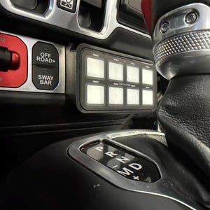 May include: A black switch panel with eight illuminated square buttons is mounted in a vehicle's dashboard. The panel is labeled "SWITCH-PRO". Adjacent are buttons labeled "OFF ROAD+" and "SWAY BAR". A gear shift lever is visible.