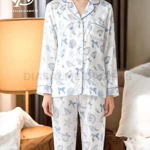 A woman wearing a white pajama top with a pattern of seashells and starfish, paired with matching pajama pants, stands in a room with a lamp and a bed visible in the background.