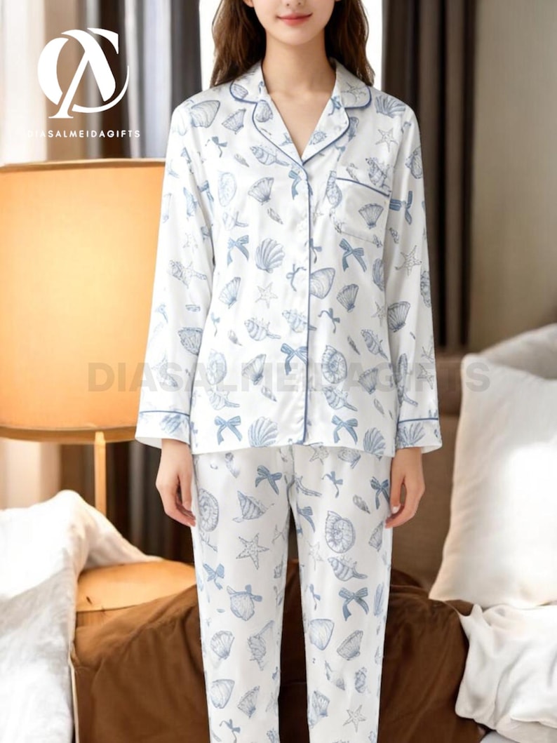 A woman wearing a white pajama top with a pattern of seashells and starfish, paired with matching pajama pants, stands in a room with a lamp and a bed visible in the background.