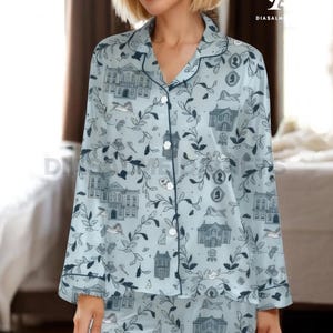 May include: Light blue pajama set with a repeating pattern of buildings, birds, and foliage. The long-sleeved top has a collared neckline and button-down front. The trim is navy blue. The pajama bottoms are visible.