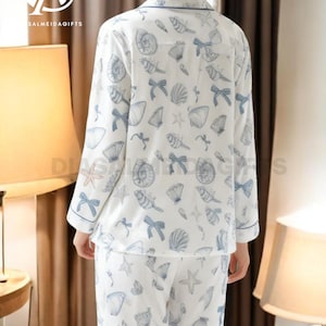 A woman wearing a white pajama top with a pattern of seashells and starfish, standing in a room with a lamp and a window.