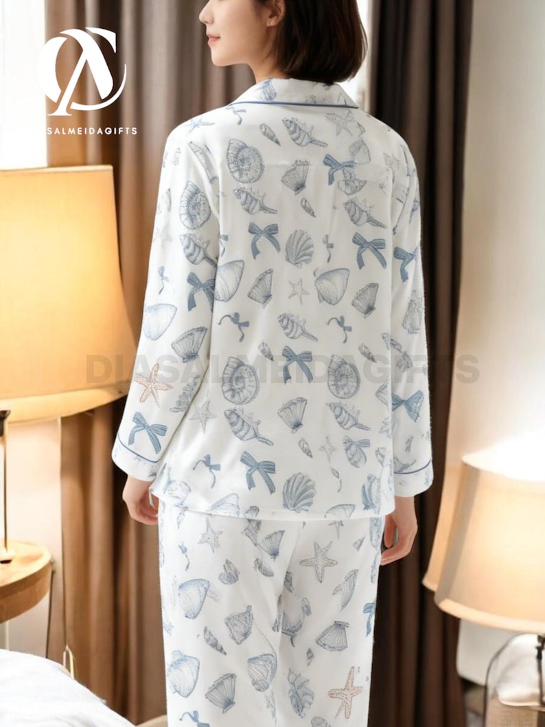 A woman wearing a white pajama top with a pattern of seashells and starfish, standing in a room with a lamp and a window.