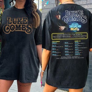 May include: Black t-shirt featuring the text "LUKE COMBS" in a brown and gold font on the front. The back of the shirt has a photo of Luke Combs and tour dates for North America and Europe. The shirt is a concert merchandise item.