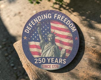 Puede incluir: Un posavasos redondo con un diseño patriótico. Presenta la Estatua de la Libertad frente a una bandera estadounidense. Las palabras "DEFENDING FREEDOM" y "250 YEARS SINCE 1776" están impresas en el posavasos.