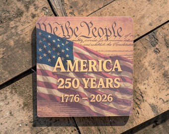Puede incluir: Cartel de madera cuadrado con la bandera americana y el texto "AMERICA 250 YEARS 1776-2026." El cartel también incluye la frase "We the People" y otras escrituras. El cartel está sobre un fondo de madera rústica.