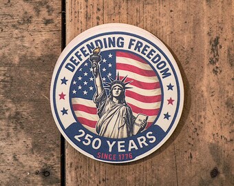 Puede incluir: Adhesivo redondo con la Estatua de la Libertad y la bandera estadounidense. El texto "Defending Freedom" y "250 Years Since 1776" se muestra. El adhesivo está sobre un fondo de madera, con una estética vintage.
