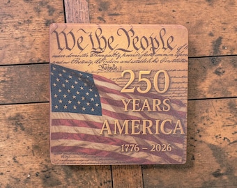 Puede incluir: Letrero cuadrado de madera con un diseño de la bandera estadounidense. El letrero presenta el texto "We the People" y "250 YEARS AMERICA 1776-2026". La bandera es roja, blanca y azul, con estrellas y rayas. El fondo tiene un aspecto de pergamino vintage.