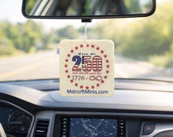 Puede incluir: Un ambientador para coche de color crema con un diseño patriótico. El diseño presenta el número 250 en rojo y azul, rodeado de estrellas rojas. También se incluye el texto "Free for 250 years and will remain free".