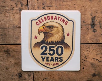 Puede incluir: Un posavasos cuadrado blanco con un diseño patriótico. El posavasos presenta una ilustración detallada de la cabeza de un águila dentro de un emblema en forma de escudo. El texto dice "Celebrating 250 Years 1776-2026".