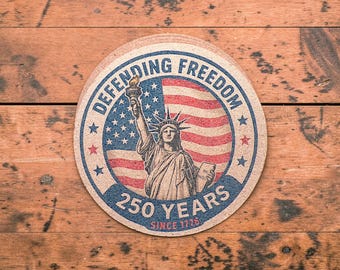 Puede incluir: Posavasos redondo con la Estatua de la Libertad frente a la bandera estadounidense. Las palabras "DEFENDING FREEDOM" y "250 YEARS SINCE 1776" están impresas. El posavasos es de material marrón claro, aprox. 10 cm de diámetro.