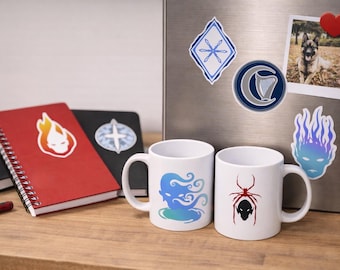 May include: Two white ceramic mugs with unique designs, one with a blue and green sea creature and the other with a red spider. A red spiral-bound notebook with a flame sticker and a black notebook are on a wooden surface.