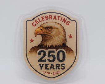 Puede incluir: Un emblema en forma de escudo con una ilustración de un águila calva. El emblema dice "CELEBRATING 250 YEARS 1776-2026" en texto rojo y negro. Dos estrellas rojas flanquean el águila. El emblema está encerrado en un borde claro y redondeado.