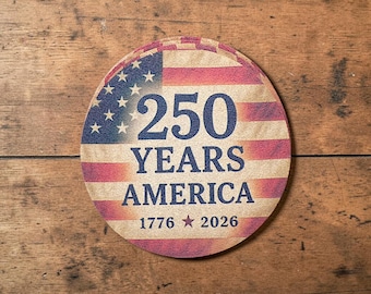 Puede incluir: Posavasos redondo con diseño de bandera estadounidense. El posavasos presenta el texto "250 YEARS AMERICA" y los años "1776 2026". El posavasos es de material marrón claro y está sobre un fondo de madera.