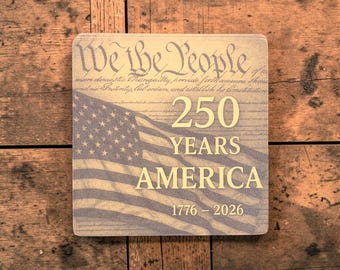 Puede incluir: Posavasos cuadrado con un diseño de la bandera estadounidense y el texto "We the People" y "250 YEARS AMERICA 1776-2026". El posavasos es de color beige y azul, sobre un fondo de madera.