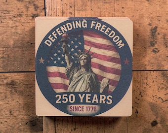 Puede incluir: Posavasos cuadrado con diseño patriótico. El posavasos presenta la Estatua de la Libertad frente a una bandera estadounidense. El texto "DEFENDING FREEDOM" forma un arco en la parte superior, con "250 YEARS SINCE 1776" en la parte inferior. Fondo de madera clara.