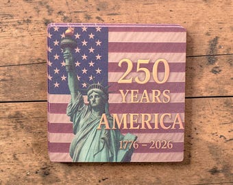 Puede incluir: Posavasos cuadrado con la Estatua de la Libertad y la bandera estadounidense. El diseño incluye el texto "250 YEARS AMERICA 1776-2026" en letras doradas. El posavasos tiene un borde rosa y es de material absorbente.