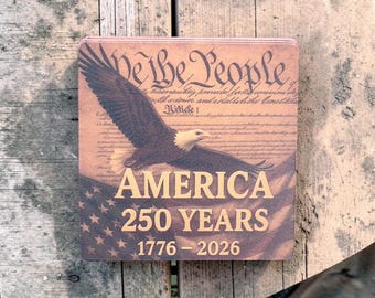 Puede incluir: Letrero cuadrado de madera con temática patriótica. Presenta un águila en vuelo sobre un diseño de bandera estadounidense. El letrero incluye el texto "We the People", "AMERICA 250 YEARS 1776-2026" y extractos de la Constitución de EE. UU.