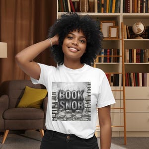 May include: White t-shirt featuring the text "BOOK SNOB" in a grayscale design. The graphic includes stacks of books and a stylized font. The shirt is worn by a person with dark curly hair.
