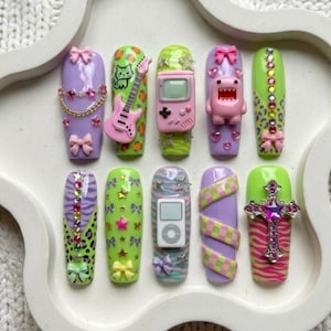 May include: A set of ten long, coffin-shaped artificial nails. Each nail features unique designs, including bows, a pink guitar, a game console, and a cross. Colors include purple, green, pink, and zebra print.