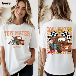 May include: Ivory-coloured t-shirt featuring a cartoon image of Tow Mater from the Cars film. The front design includes the text "TOW MATER EST. 2006 CARS" with a checkered flag. The back design shows Mater with checkered flags and the text "PISTON CUP ROUTE 66".