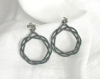 May include: A pair of silver-toned hoop earrings. Each earring features a woven design, creating a textured, circular shape. The earrings are displayed on a white fabric background, highlighting their metallic sheen and intricate details. These earrings are a stylish accessory.