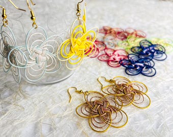 May include: Floral-shaped earrings in various colors, including yellow, blue, and gold, with gold-colored ear wires. Several pairs are displayed, with some in a clear glass container. A collection of colorful earrings is also visible.