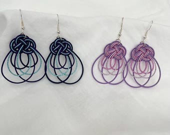 May include: Two pairs of handmade earrings. One pair features a dark blue knot with teal and black accents, and the other pair has a purple knot with pink and silver accents. Both pairs have a teardrop shape with multiple loops.