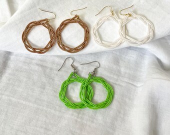 May include: Four pairs of hoop earrings in various colors are displayed on a white cloth. The earrings include two pairs of bronze-colored hoops, two pairs of white hoops, and a pair of bright green hoops. All earrings have a woven design.