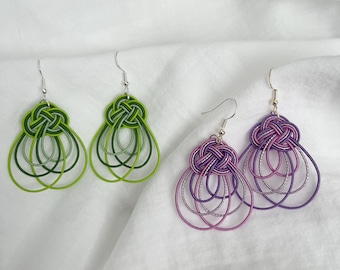 May include: Two pairs of handmade earrings. One pair is green, with a knot design at the top and layered loops below. The other pair is purple, with a similar knot design and layered loops. The earrings hang from silver hooks.