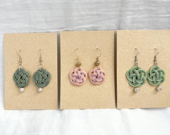 May include: Three pairs of handmade earrings displayed on tan cards. The earrings feature intricate knotwork in green, pink, and green with silver or gold accents. Each pair has a unique design and color combination, showcasing the craftsmanship.