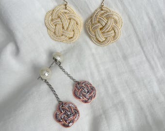 May include: Two pairs of earrings on a white surface. One pair features gold-toned hooks and cream-colored woven designs. The other pair has pearl studs, silver chains, and rose gold and silver woven flower-shaped pendants.