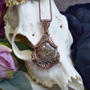 May include: A handcrafted copper wire pendant necklace. The pendant features a round, speckled stone in shades of brown and gray, encased in intricate copper wirework. The necklace has a delicate chain and is displayed against a skull.