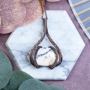 Op de afbeelding: Een handgemaakte hangerketting met een witte en grijze steen gezet in een koperdraadframe. De hanger hangt aan een fijne koperen ketting. De steen heeft een kenmerkend patroon en het draadwerk geeft de sieraden een artistieke uitstraling.