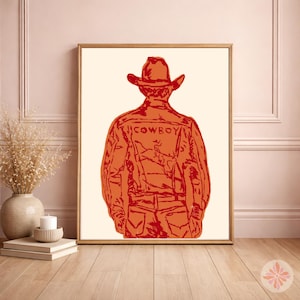 May include: A framed wall art print featuring a stylized orange and red line drawing of a person wearing a cowboy hat and a jacket with the word COWBOY printed across the back. The illustration is set against a plain off-white background.
