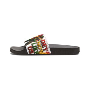 May include: Black slide sandal with a white strap featuring a colorful graphic and the text "DEY NOT LIKE US". The graphic includes a cartoon character in green, red, and yellow.