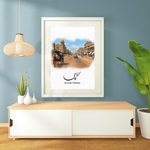May include: Framed art print depicting a street scene in Karachi, Pakistan. The artwork features a horse-drawn carriage, buildings, and the city's name in Urdu script. The print is displayed on a white frame, set against a blue wall.