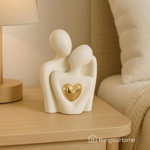 May include: A white ceramic sculpture of two embracing figures, featuring a gold heart. The figures have a smooth, minimalist design. The sculpture is placed on a wooden surface. The overall aesthetic is modern and elegant.