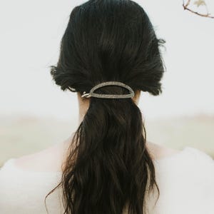 May include: A decorative hair clip with a silver finish, set with small, sparkling stones, is placed in dark hair styled in a low ponytail. The clip is oval-shaped and adds a touch of elegance to the hairstyle. The background is blurred.