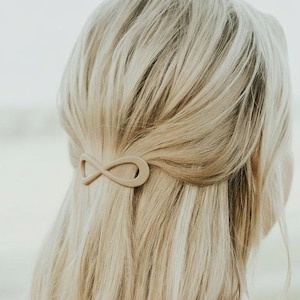 May include: A light brown infinity hair clip is holding back blonde hair. The hair clip is made of a matte material and is in the shape of an infinity symbol. The hair is styled in a half-up, half-down style.