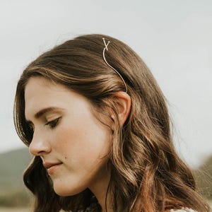 May include: A woman with long, wavy brown hair wearing a white lace top and a delicate gold hair accessory. The background is a field with a cloudy sky and distant hills.