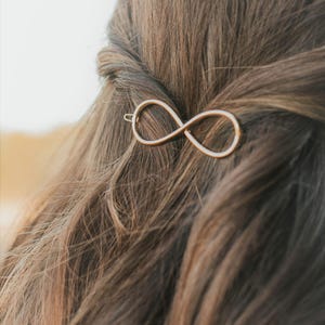 May include: A gold-colored hair clip in the shape of an infinity symbol is pinned into brown hair. The clip is made of a thin metal and is positioned on the side of the head, holding a section of hair in place.
