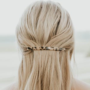 May include: A rectangular hair barrette featuring a beige base with a black and dark brown tortoiseshell pattern. The hair accessory is positioned horizontally, securing a section of blonde hair at the back of the head in a half-up style.